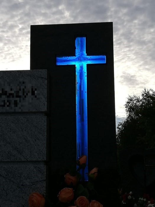 Niebiesko-granatowy krzyż ze szkła cieniowany od najjaśniejszego szkła na brzegach do najciemniejszego w środku. Krzyż wmontowany w czarną płytę, prześwietlony słońcem wygląda jakby świecił własnym światłem.