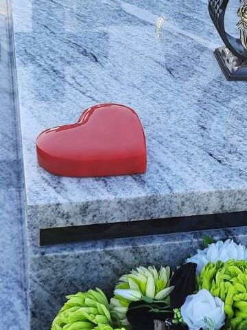 czerwone niewielkie serduszko przyklejone do  szarej płyty poziomej nagrobka po lewej stronie pod skosem. na środku nagrobka stoją znicze a z przodu kompozycja z biało-zielonych duzych chryzantem.